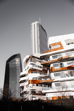 A cityscape featuring modern architecture with two prominent buildings. One is a sleek, dark tower with the PwC logo, while the other is a white building with wooden accents and multiple balconies. The design is contemporary, with rounded edges and a mix of materials, creating an urban, sophisticated atmosphere. Leafless trees are visible in the foreground.