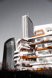 A cityscape featuring modern architecture with two prominent buildings. One is a sleek, dark tower with the PwC logo, while the other is a white building with wooden accents and multiple balconies. The design is contemporary, with rounded edges and a mix of materials, creating an urban, sophisticated atmosphere. Leafless trees are visible in the foreground.