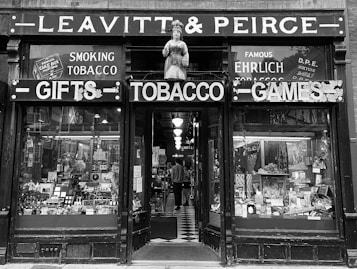 A vintage storefront with a figure statue above the entrance, showcasing signs for gifts, tobacco, and games. The display windows are filled with an assortment of products including tobacco items. Inside, the shop is dimly lit with visible shelves and a checkered floor leading inwards. The facade gives an antique and historical feel.