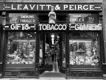 A vintage storefront with a figure statue above the entrance, showcasing signs for gifts, tobacco, and games. The display windows are filled with an assortment of products including tobacco items. Inside, the shop is dimly lit with visible shelves and a checkered floor leading inwards. The facade gives an antique and historical feel.
