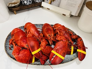 A platter holds multiple boiled lobsters with rubber bands on their claws resting on a countertop. The background includes a cookbook stand and some kitchen items.