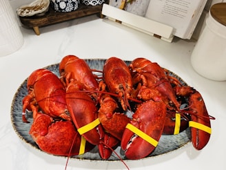 A platter holds multiple boiled lobsters with rubber bands on their claws resting on a countertop. The background includes a cookbook stand and some kitchen items.