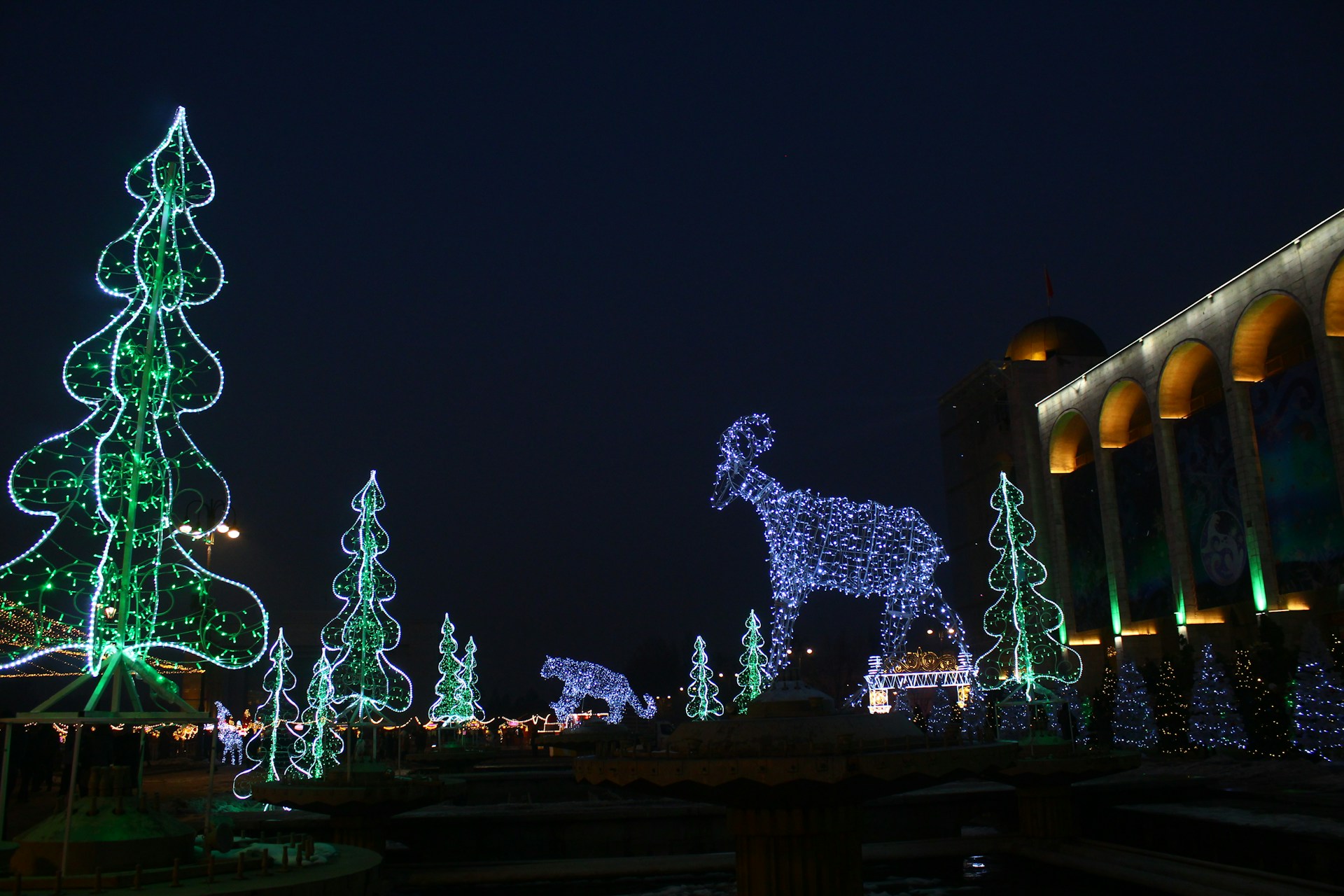 A nighttime scene featuring large illuminated decorations shaped like trees and animals, including a prominent reindeer figure made of blue lights. The area is adorned with several luminous green tree shapes. A building with lit arches is visible on the right side.
