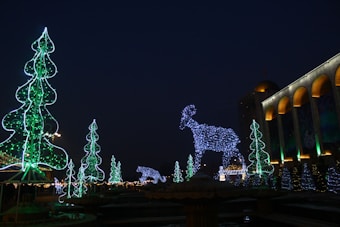 A nighttime scene featuring large illuminated decorations shaped like trees and animals, including a prominent reindeer figure made of blue lights. The area is adorned with several luminous green tree shapes. A building with lit arches is visible on the right side.