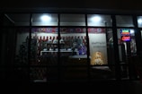 A dimly lit store showcasing a variety of traditional items and decorations. The window display includes colorful patterned textiles, stuffed toys, and decorative ornaments. There is a small tree and illuminated signage with Cyrillic text, indicating the shop is open.