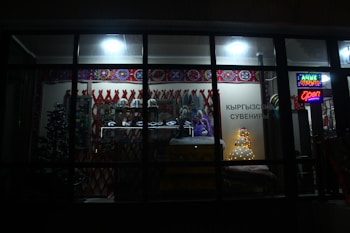 A dimly lit store showcasing a variety of traditional items and decorations. The window display includes colorful patterned textiles, stuffed toys, and decorative ornaments. There is a small tree and illuminated signage with Cyrillic text, indicating the shop is open.