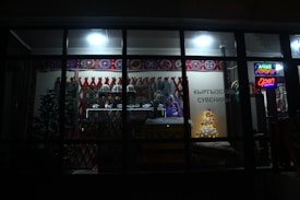 A dimly lit store showcasing a variety of traditional items and decorations. The window display includes colorful patterned textiles, stuffed toys, and decorative ornaments. There is a small tree and illuminated signage with Cyrillic text, indicating the shop is open.