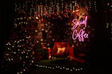 A cozy Christmas-themed setting with a decorated tree adorned with lights and ornaments. The backdrop features a wall with snowflake patterns and a neon 'Social Coffee' sign in pink. Under the decorations, there's a chair with a festive pillow, surrounded by wrapped gifts and a red Santa hat, all illuminated by warm lighting.