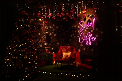 A cozy Christmas-themed setting with a decorated tree adorned with lights and ornaments. The backdrop features a wall with snowflake patterns and a neon 'Social Coffee' sign in pink. Under the decorations, there's a chair with a festive pillow, surrounded by wrapped gifts and a red Santa hat, all illuminated by warm lighting.