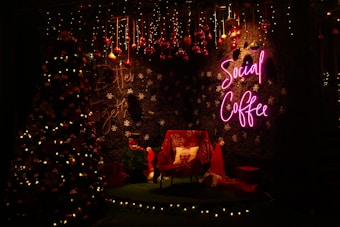 A cozy Christmas-themed setting with a decorated tree adorned with lights and ornaments. The backdrop features a wall with snowflake patterns and a neon 'Social Coffee' sign in pink. Under the decorations, there's a chair with a festive pillow, surrounded by wrapped gifts and a red Santa hat, all illuminated by warm lighting.