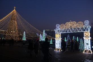 A large Christmas tree adorned with golden lights dominates the scene, with a glowing archway decorated in intricate patterns on the right. Several smaller illuminated trees and festive decorations are scattered in the background. People are walking around, enjoying the festive atmosphere during twilight.