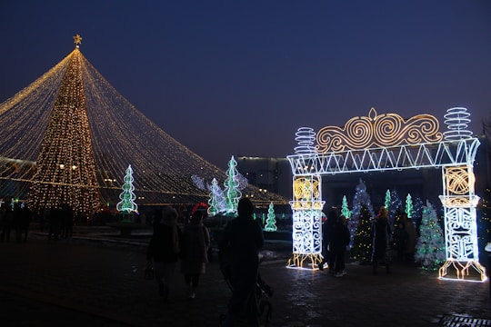 A large Christmas tree adorned with golden lights dominates the scene, with a glowing archway decorated in intricate patterns on the right. Several smaller illuminated trees and festive decorations are scattered in the background. People are walking around, enjoying the festive atmosphere during twilight.
