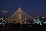 A well-lit Christmas tree with strings of lights radiating upward, surrounded by people and other decorative trees with green and blue lights. The scene takes place during the evening, with street lamps casting a warm glow over the area.