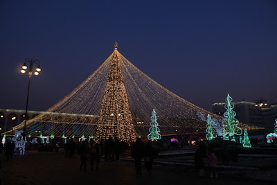 A well-lit Christmas tree with strings of lights radiating upward, surrounded by people and other decorative trees with green and blue lights. The scene takes place during the evening, with street lamps casting a warm glow over the area.