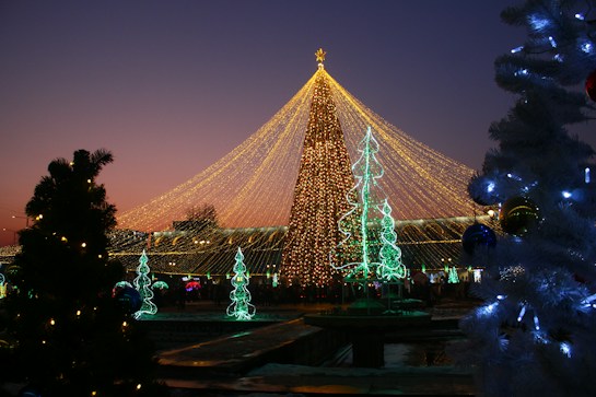 A festive scene featuring a large Christmas tree adorned with golden lights, forming an intricate canopy above a public space. Smaller trees are outlined with green lights, contributing to the celebratory atmosphere. The image includes people walking around, illuminated ornaments hanging from trees, and multi-colored lights creating a vibrant and joyful environment. The sky in the background showcases a gradient from deep blue to soft pink, enhancing the visual appeal.