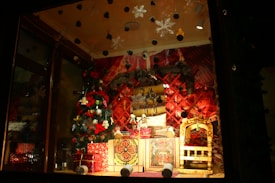A festive window display featuring a decorated Christmas tree with ornaments and lights, surrounded by red-wrapped gift boxes. The backdrop consists of a red and gold patterned wall with hanging decorations and a plush animal figure. A small wooden chair with intricate carvings is also present.