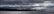 Dark clouds cover the sky over a large body of water, with distant mountains partially visible under the heavy cloud cover. The atmosphere appears moody and dramatic as light attempts to break through in certain areas, creating a serene yet ominous ambiance.