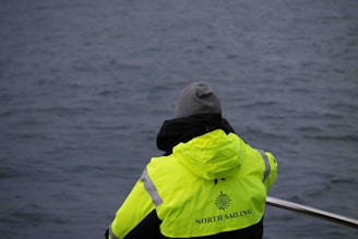 A person wearing a bright yellow jacket with 'North Sailing' written on it is standing by the railing, gazing at the calm blue sea. The person is wearing a gray hood underneath the jacket.