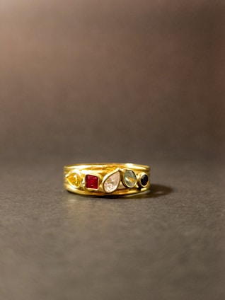 A gold ring adorned with multiple gemstones sits on a flat surface. The stones come in various shapes and colors, including red, blue, green, and clear, set within the band