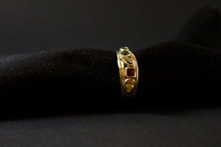 A gold ring adorned with various gemstones is displayed against a soft, black textured background. The gemstones include red, blue, and transparent stones, each embedded in the band, adding vibrant colors to the ring's elegant design.
