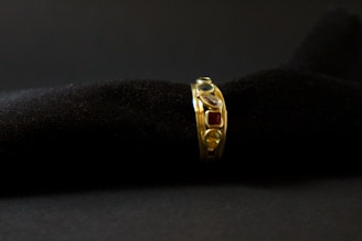 A gold ring adorned with various gemstones is displayed against a soft, black textured background. The gemstones include red, blue, and transparent stones, each embedded in the band, adding vibrant colors to the ring's elegant design.