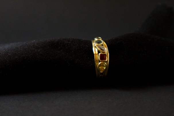 A gold ring adorned with various gemstones is displayed against a soft, black textured background. The gemstones include red, blue, and transparent stones, each embedded in the band, adding vibrant colors to the ring's elegant design.
