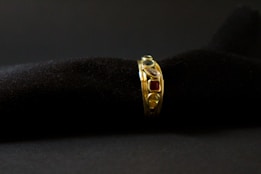 A gold ring adorned with various gemstones is displayed against a soft, black textured background. The gemstones include red, blue, and transparent stones, each embedded in the band, adding vibrant colors to the ring's elegant design.
