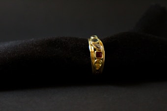 A gold ring adorned with various gemstones is displayed against a soft, black textured background. The gemstones include red, blue, and transparent stones, each embedded in the band, adding vibrant colors to the ring's elegant design.