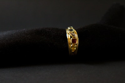 A gold ring adorned with various gemstones is displayed against a soft, black textured background. The gemstones include red, blue, and transparent stones, each embedded in the band, adding vibrant colors to the ring's elegant design.