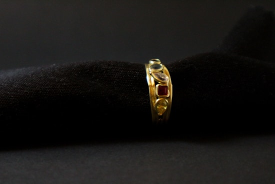 A gold ring adorned with various gemstones is displayed against a soft, black textured background. The gemstones include red, blue, and transparent stones, each embedded in the band, adding vibrant colors to the ring's elegant design.