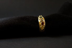 A gold ring adorned with various gemstones is displayed against a soft, black textured background. The gemstones include red, blue, and transparent stones, each embedded in the band, adding vibrant colors to the ring's elegant design.