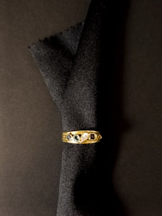A gold ring with five distinct gemstones rests against a dark fabric background. The gems include a blue stone, a green stone, a clear stone, and a red stone, along with another smaller dark stone.