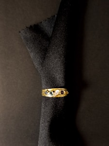 A gold ring with five distinct gemstones rests against a dark fabric background. The gems include a blue stone, a green stone, a clear stone, and a red stone, along with another smaller dark stone.