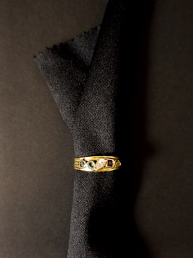 A gold ring with five distinct gemstones rests against a dark fabric background. The gems include a blue stone, a green stone, a clear stone, and a red stone, along with another smaller dark stone.