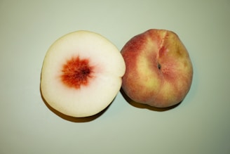 Two peaches are placed on a light green surface. One peach is whole with a velvety skin featuring shades of pink, red, and yellow, while the other is cut in half displaying its light cream flesh with a deep red pit in the center.