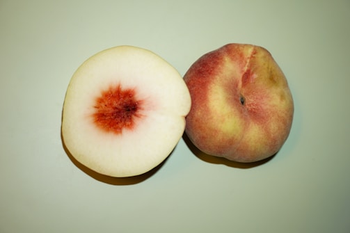 Two peaches are placed on a light green surface. One peach is whole with a velvety skin featuring shades of pink, red, and yellow, while the other is cut in half displaying its light cream flesh with a deep red pit in the center.