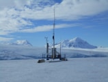 A remote weather station is situated in a vast, snowy landscape. Majestic snow-covered mountains rise in the background under a partly cloudy sky. The station is equipped with various antennas and instruments, perched on a flat expanse of ice, with a small red flag marking the area.