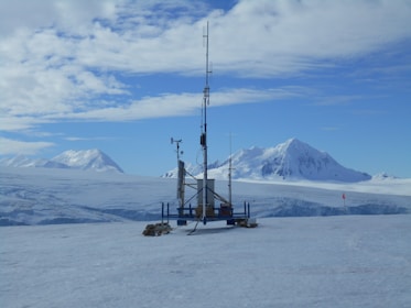 A remote weather station is situated in a vast, snowy landscape. Majestic snow-covered mountains rise in the background under a partly cloudy sky. The station is equipped with various antennas and instruments, perched on a flat expanse of ice, with a small red flag marking the area.