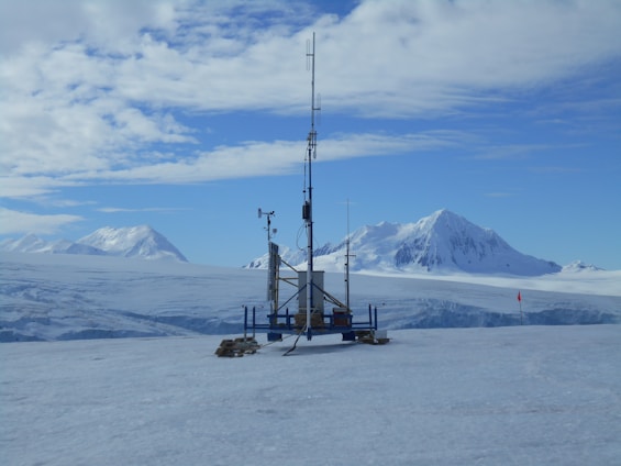 A remote weather station is situated in a vast, snowy landscape. Majestic snow-covered mountains rise in the background under a partly cloudy sky. The station is equipped with various antennas and instruments, perched on a flat expanse of ice, with a small red flag marking the area.