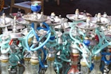 An array of hookahs with various colored hoses and metal bowls. The hookahs are tightly packed together, showcasing a mix of metallic finishes and vibrant hues. The shimmering highlights on the metal parts contrast with the colorful, flexible hoses that wind around each other.