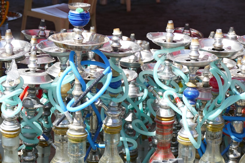 An array of hookahs with various colored hoses and metal bowls. The hookahs are tightly packed together, showcasing a mix of metallic finishes and vibrant hues. The shimmering highlights on the metal parts contrast with the colorful, flexible hoses that wind around each other.