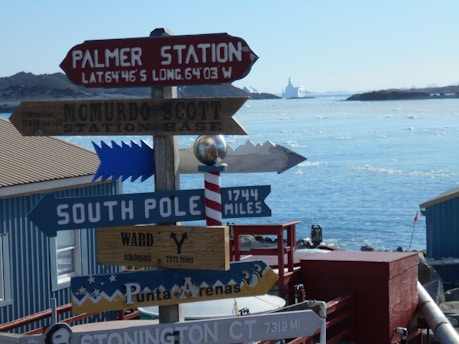 Several directional signs are pointing towards various locations such as Palmer Station, McMurdo, Scott Station Base, South Pole, Ward, and Punta Arenas. The signs indicate the distances in miles to these places. In the background, there is a view of an icy landscape with calm water and icebergs, under a clear blue sky.