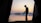 A silhouette of a person holding a camera is framed by the open flaps of a tent. The background features a calm lake reflecting the colors of the sunset, with distant hills and a sky transitioning from orange to blue.