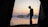 A silhouette of a person holding a camera is framed by the open flaps of a tent. The background features a calm lake reflecting the colors of the sunset, with distant hills and a sky transitioning from orange to blue.