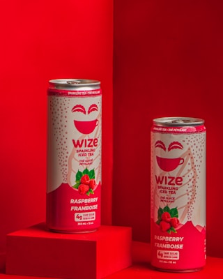 Two cans of Wize Raspberry Framboise Sparkling Iced Tea are positioned against a vibrant red background, one on a red pedestal and the other slightly behind. The design on the cans includes an illustration of raspberries and a cup, along with text indicating the flavor and brand.