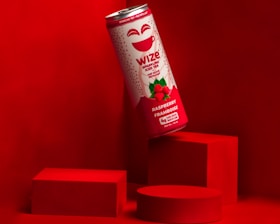 A can of Wize Raspberry Iced Tea is prominently displayed against a vibrant red background with geometric shapes. The can features a white and red color scheme with green raspberry illustrations.