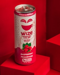 A can of Wize sparkling iced tea with raspberry flavor is placed against a red geometric background. The can features a design with raspberry illustrations and the label includes both English and French text. The metallic top of the can is visible.