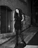Black and white photo of a model wearing a velvet goth dress with lace details, standing against a cracked urban wall.