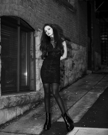 Black and white photo of a model wearing a velvet goth dress with lace details, standing against a cracked urban wall.