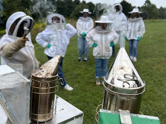 A group of people wearing protective beekeeping suits with veils stand in a grassy field. Two metal bee smokers are in the foreground, emitting smoke. The individuals are dressed uniformly, with white suits and green gloves, and trees are visible in the background.